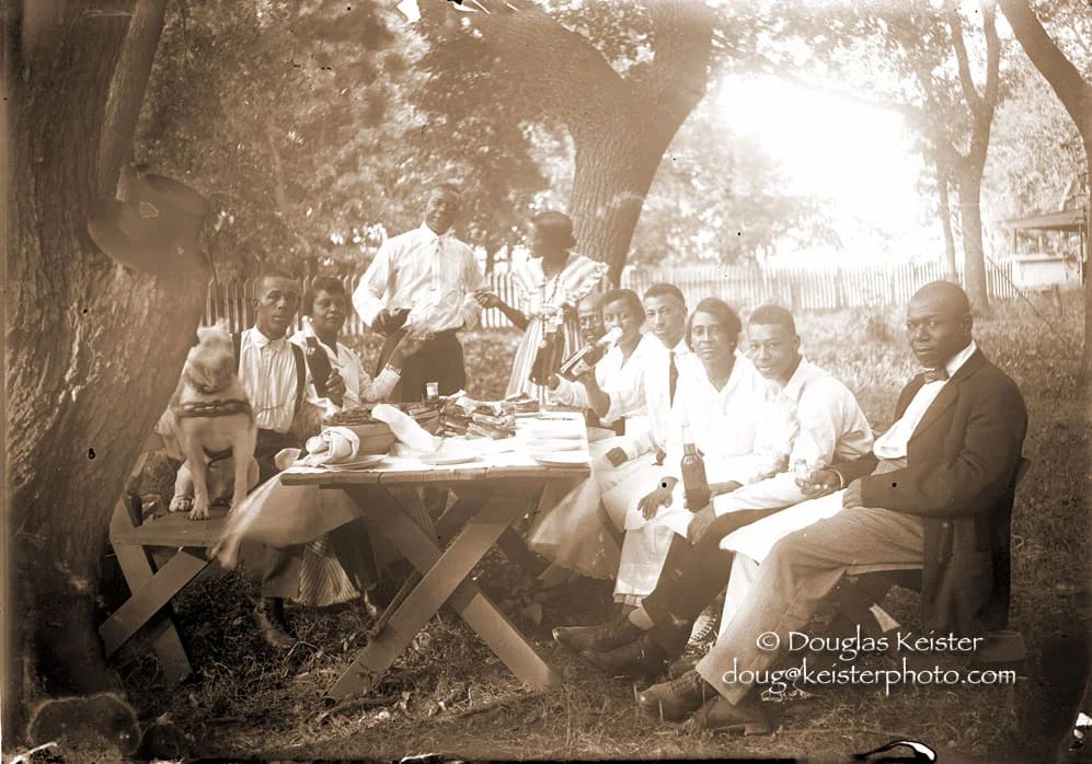Group of men and women dining outdoors under large trees, circa early 20th century.
