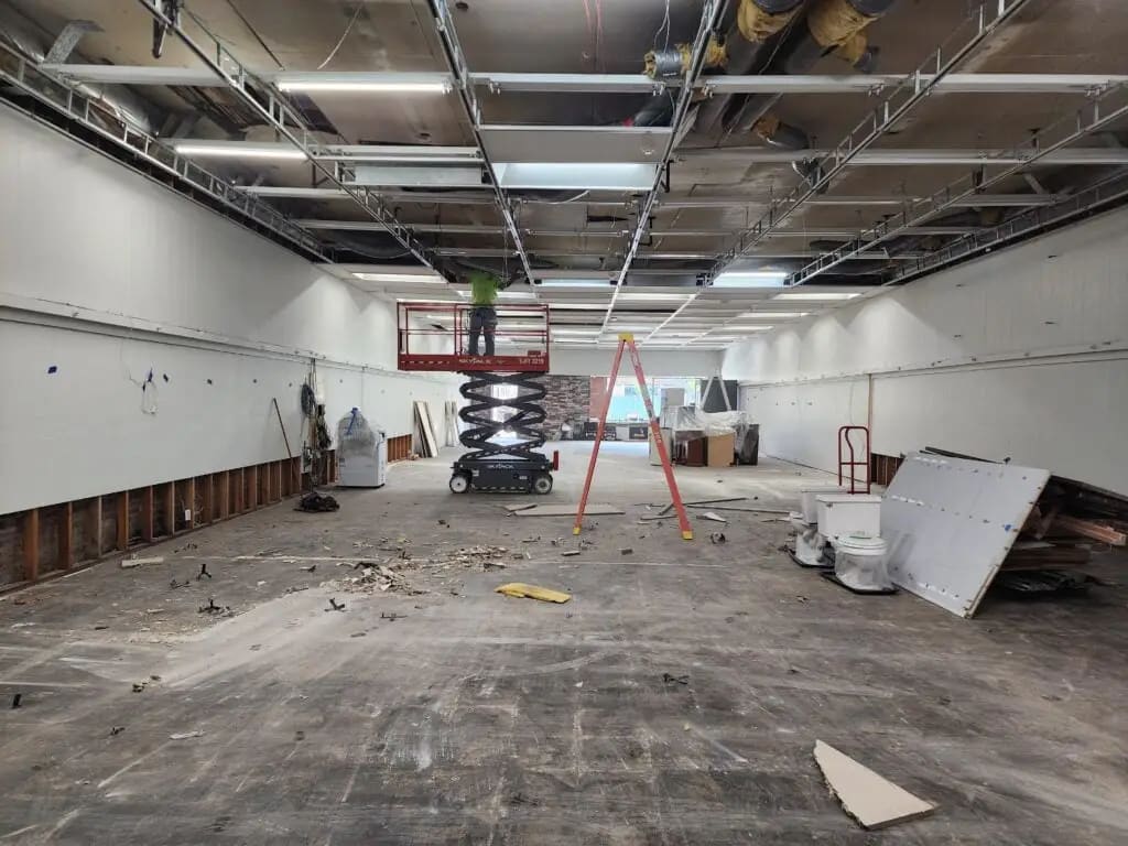 Interior construction site with scissor lift, ladders, and debris in an empty commercial space.