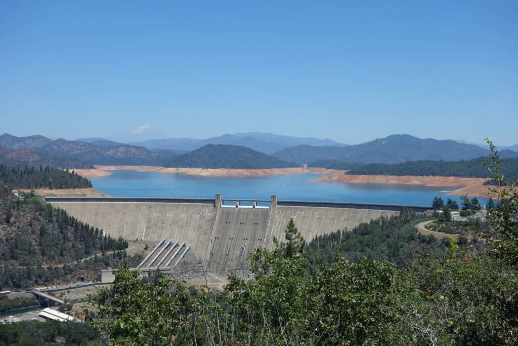 Large concrete dam holding back a blue reservoir, surrounded by forested mountains under clear sky.