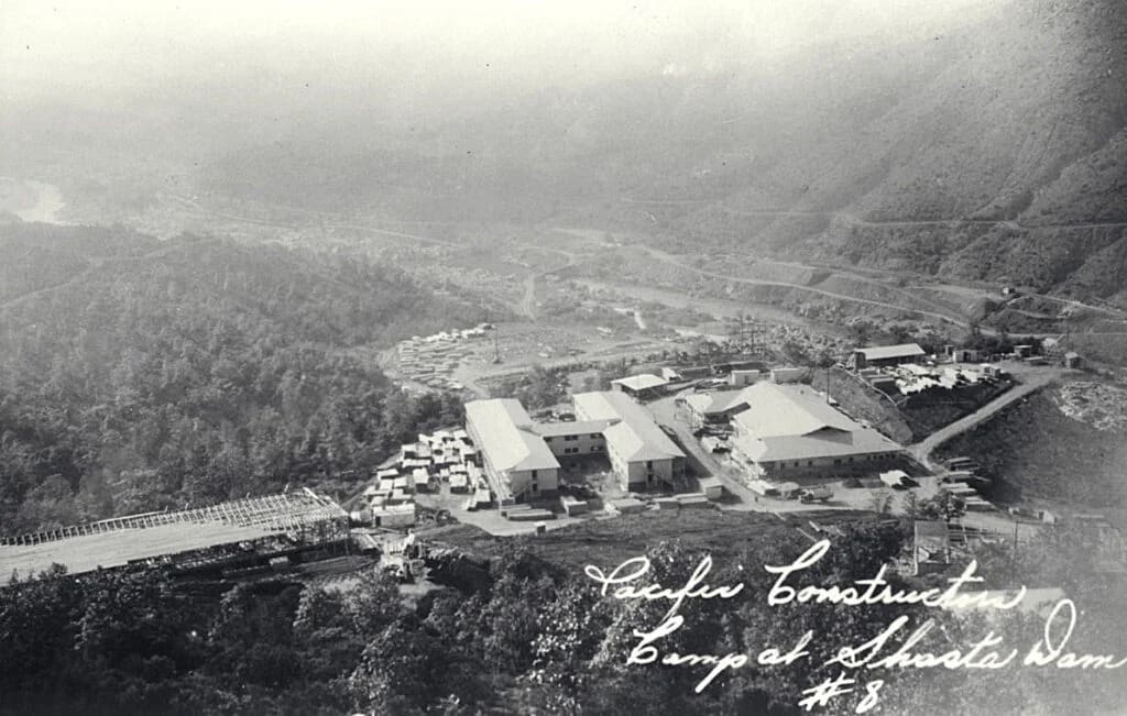 Aerial view of large industrial or institutional buildings with white roofs nestled in a forested mountain valley with winding roads.