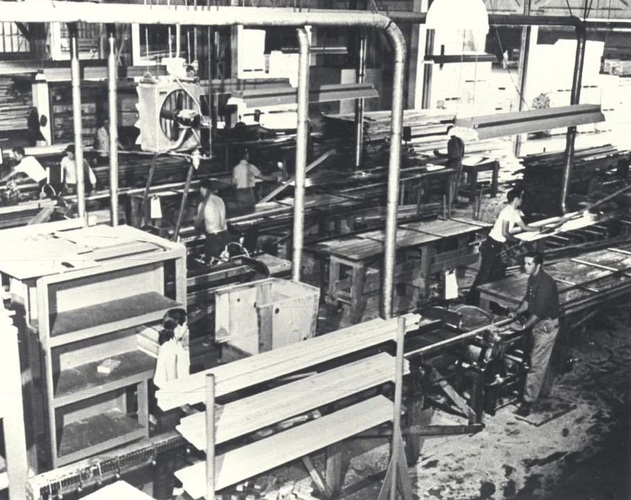 Workers operate machinery in a mid-century woodworking or furniture factory workshop.