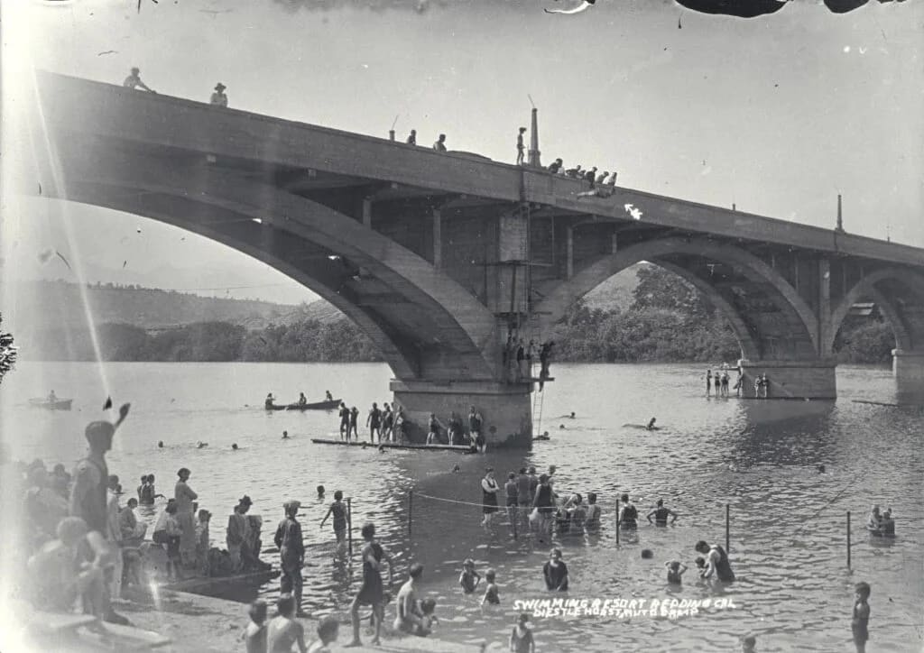 Large crowds of swimmers and spectators enjoy a swimming and sport outing under a concrete bridge spanning a river.