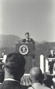 Official speaking at a podium with presidential seal before an outdoor crowd.